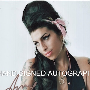 May include: A photographic print featuring Amy Winehouse with her signature beehive hairstyle, a pink hair ribbon, and a black sleeveless polo shirt. The image includes a hand-signed autograph in brown ink and the text HAND SIGNED AUTOGRAPH.