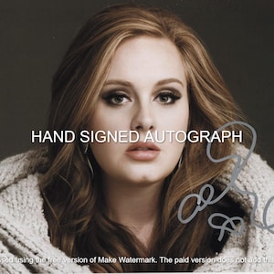 Puede incluir: Un retrato de una mujer con cabello largo y ondulado castaño, vistiendo un suéter de color claro. La imagen incluye el texto "HAND SIGNED AUTOGRAPH" y una firma. El fondo es de un color neutro sólido.