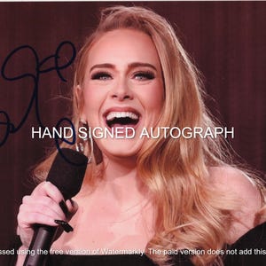 Puede incluir: Fotografía de una mujer sonriente sosteniendo un micrófono. La imagen incluye el texto "HAND SIGNED AUTOGRAPH" y una firma manuscrita. La mujer tiene el pelo largo y rubio ondulado y lleva una blusa negra.