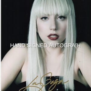 May include: A portrait of Lady Gaga with long, straight platinum blonde hair and bangs, wearing a black top. The image features the text HAND SIGNED AUTOGRAPH in white and a gold-colored signature at the bottom against a dark background.