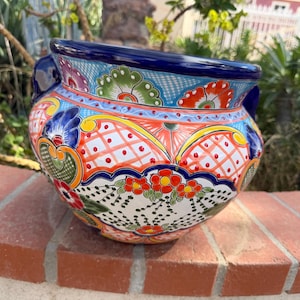 May include: A round ceramic planter featuring a vibrant hand-painted design with a deep blue rim and handles. The surface is decorated with intricate floral patterns, geometric grids, and scalloped motifs in bright red, orange, green, and yellow.