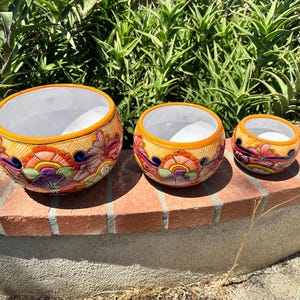 May include: Three decorative ceramic bowls with a vibrant floral design. The bowls have a white interior, a yellow rim, and a colorful exterior with orange, blue, purple, and green floral patterns. The bowls are arranged in a row, decreasing in size.