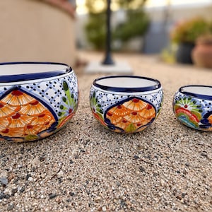 May include: A set of three round ceramic bowls featuring a hand-painted pattern of orange fan-shaped motifs, green accents, and a blue grid design on a white background, with a dark blue rim around the top edge of each bowl.