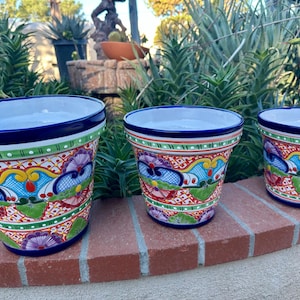 May include: Three ceramic flower pots with a vibrant, hand-painted design. The pots feature a blue rim, a white interior, and a colorful exterior with floral and geometric patterns in red, green, yellow, and blue. They are arranged on a brick surface.