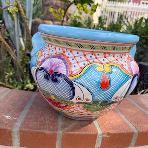 May include: A round ceramic planter featuring a light blue rim and handles, decorated with a vibrant, multi-colored pattern of red, blue, and purple floral motifs, intricate cross-hatching, and geometric shapes on a white background.