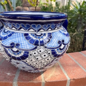 May include: A round ceramic planter featuring a glossy finish with intricate hand-painted patterns in deep blue, light blue, and white. The design includes floral motifs, checkered sections, and dotted grids, complemented by two small side handles.