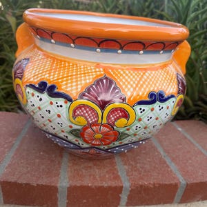 May include: A round ceramic planter featuring a vibrant orange rim and two side handles. The body is decorated with a colorful pattern of red flowers, yellow scrolls, and green cross-hatching on a white background with blue and purple accents.