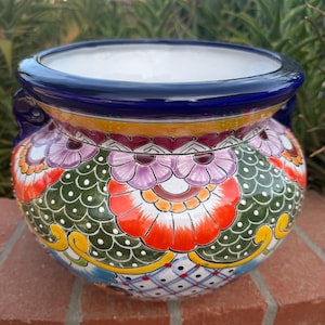 May include: A round ceramic planter featuring a wide, dark blue rim and handles. The exterior is decorated with a vibrant, multi-colored pattern of orange, green, and purple floral motifs, along with a white grid design containing small red dots.