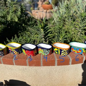 May include: Seven oval ceramic planters arranged in a row, each featuring distinct hand-painted Talavera-style patterns in vibrant colors including blue, red, yellow, and green. Each planter is numbered one through seven in blue ink.