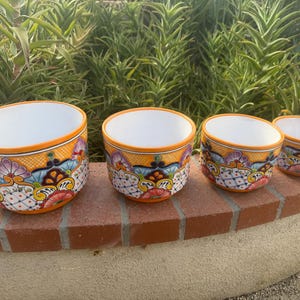 May include: A set of four round ceramic planters in graduated sizes, each featuring a vibrant hand-painted pattern with orange rims, white interiors, and intricate floral and geometric motifs in shades of red, blue, purple, and yellow.