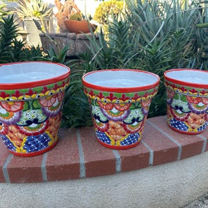 May include: Three colorful ceramic flower pots with a white interior, set on a brick ledge. The pots feature a vibrant design with red, green, orange, blue, and purple floral patterns. The rims are red, and the pots vary in size.
