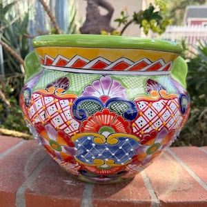 May include: A round ceramic planter featuring a vibrant hand-painted design with a lime green rim, orange geometric patterns, and intricate floral motifs in shades of red, blue, purple, and yellow. The pot includes two side handles.