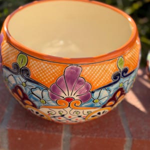 May include: A round ceramic planter featuring a vibrant hand-painted design with orange, purple, light blue, and dark blue floral and geometric patterns. The interior is a solid cream color, and the rim is finished with a bright orange glaze.