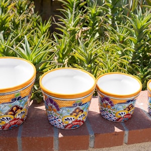 May include: Four ceramic flower pots with a white interior and a colorful exterior design. The pots feature a yellow rim and a pattern of flowers and geometric shapes in blue, red, and orange. They are arranged in a row on a brick surface.