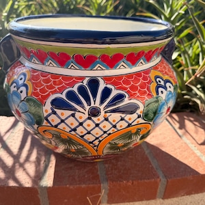 May include: A round ceramic planter featuring a vibrant hand-painted design with a dark blue rim and handles. The body displays a colorful pattern of red, orange, yellow, and blue floral motifs, geometric shapes, and scalloped details throughout.