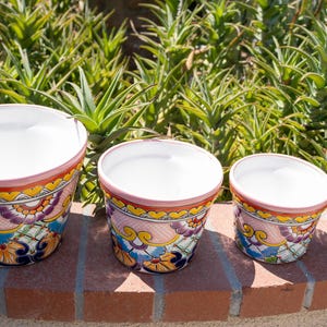 May include: Four ceramic flower pots with a white interior and a colorful exterior design. The pots have a pink rim and a decorative pattern of flowers and leaves in shades of yellow, blue, and purple. They are arranged in a row on a brick surface.