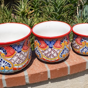 May include: Three ceramic flower pots with a white interior and a red rim. The exterior features a colorful, intricate design with blue, yellow, and red patterns. The pots are of varying sizes and sit on a brick surface.