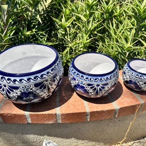 May include: Three ceramic planters with a white interior and a blue and white floral design. The planters are arranged in a row, decreasing in size from left to right. The planters sit on a brick surface with green foliage in the background.