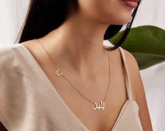 May include: A gold-colored necklace with two name pendants in Arabic script. The necklace is worn around the neck and rests on a person's chest. The pendants are connected by a delicate chain.