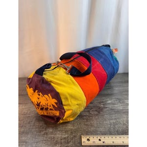 May include: A colorful duffel bag with a rainbow design. The bag features a variety of colors, including blue, red, orange, yellow, and purple. The bag has a black handle and a silver-colored metal clasp. The text "Boca Raton" is printed on the side.