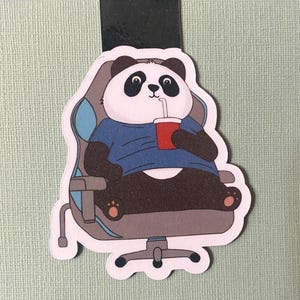 May include: A die-cut panda bear sticker, seated in a grey and blue gaming chair, holding a red cup with a straw. The panda is wearing a blue shirt. The sticker has a black magnetic strip on the top.