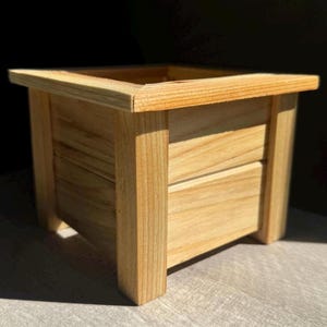 May include: A square wooden planter box with a natural wood finish. The planter has a simple design with a smooth surface and straight edges.