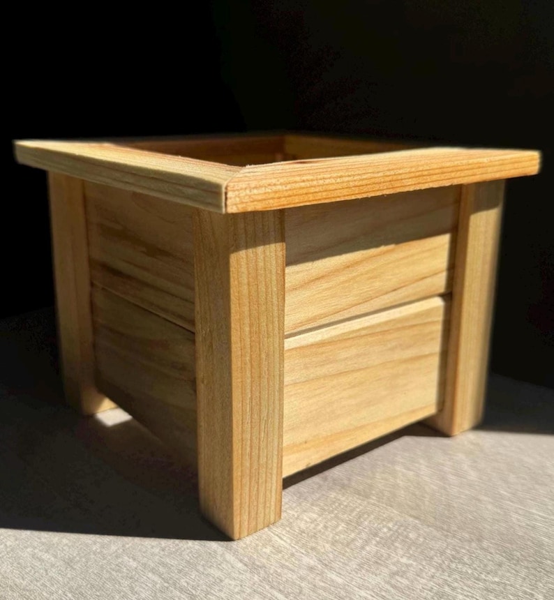 May include: A square wooden planter box with a natural wood finish. The planter has a simple design with a smooth surface and straight edges.