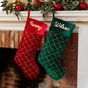 May include: Two Christmas stockings, one red and one green, hang from a fireplace mantel. The red stocking has the name "Owerry" in gold script, and the green stocking has the name "Wilson". Both stockings have a quilted pattern.