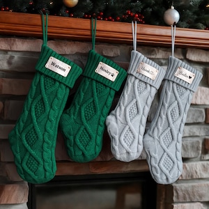 May include: Four cable-knit Christmas stockings in green and grey hang from a mantelpiece. Each stocking has a personalised tag with a name. The green stockings have the names "Watson" and "Hosley". The grey stockings have the names "Uriah" and "Bailen".