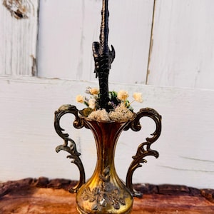 May include: A gold decorative vase with ornate handles and a black bird's foot on top. The vase is filled with dried flowers and moss.