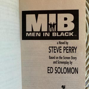 Peut inclure: Livre ouvert pr&eacute;sentant la couverture du roman &laquo; Men in Black &raquo;. Le titre est en noir, avec deux silhouettes. Le nom de l'auteur, Steve Perry, et Ed Solomon sont &eacute;galement cr&eacute;dit&eacute;s.