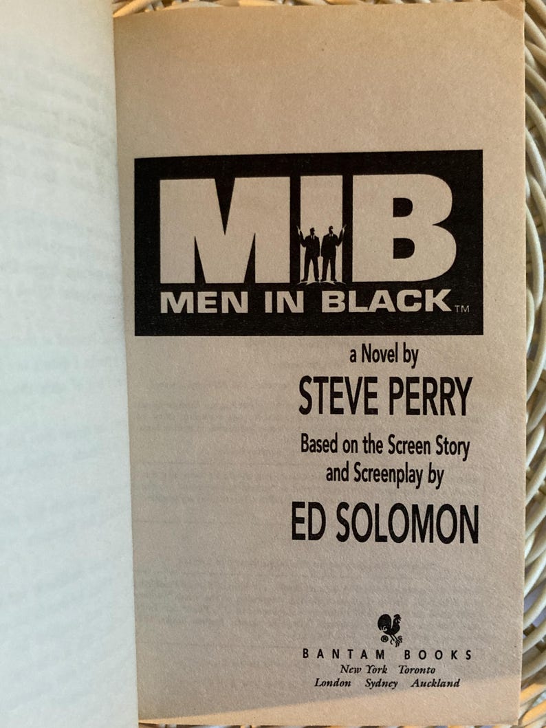 Peut inclure: Livre ouvert pr&eacute;sentant la couverture du roman &laquo; Men in Black &raquo;. Le titre est en noir, avec deux silhouettes. Le nom de l'auteur, Steve Perry, et Ed Solomon sont &eacute;galement cr&eacute;dit&eacute;s.