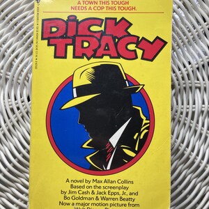 May include: A vintage paperback book titled "Dick Tracy" with a yellow cover. The title is in large red letters. The cover features a silhouette of a man in a fedora and trench coat. The book is a novel based on the screenplay.