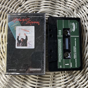 May include: A vintage cassette tape of "Midnight Express" soundtrack. The cover features a movie still with the title in red and a passport graphic. The cassette is black with a green label and the text "The Original Motion Picture Soundtrack Midnight Express".