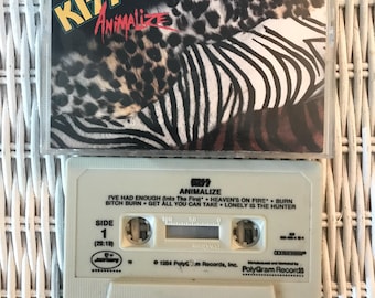 May include: A vintage cassette tape of the KISS album "Animalize." The cassette case features a cover with a zebra and leopard print design. The cassette itself is off-white with the album title and tracklist printed on it. The album was released in 1984.