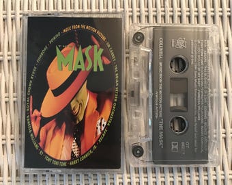 May include: Two clear cassette tapes. One has a cover with the word "MASK" in green, and an image of a man in a yellow hat and orange suit. The other tape is clear with the text "COLUMBIA MUSIC FROM THE MOTION PICTURE 'THE MASK'" printed on it.