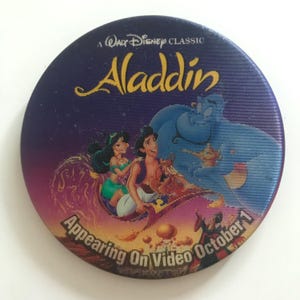 May include: A purple and blue round button with a picture of Aladdin, Jasmine, and Genie from the Disney movie Aladdin. The text on the button reads "A Walt Disney Classic Aladdin Appearing On Video October 1".
