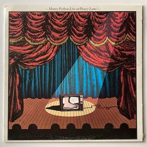 May include: A square vinyl record cover titled Monty Python Live at Drury Lane. The illustration depicts a stage with red curtains, a wooden floor, and a spotlight shining on a small television set displaying a human foot on its screen.