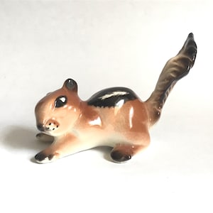 May include: A ceramic chipmunk figurine in a resting pose. The chipmunk has a brown and tan body with a black and white stripe down its back. The figurine has black eyes and a long, curved tail.