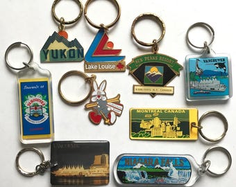 May include: A collection of ten keychains featuring various Canadian landmarks and cities. The keychains are made of metal and plastic and feature colorful designs. Some of the keychains include the text "YUKON", "Lake Louise", "SUN PEAKS RESORT", "VANCOUVER", "Souvenir of VANCOUVER", "MONTREAL CANADA", and "NIAGARA FALLS".