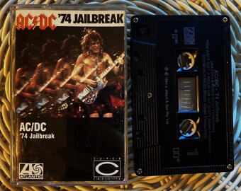 May include: A vintage AC/DC '74 Jailbreak cassette tape. The cover features a live performance image of the band. The cassette is black with white text. The Atlantic Records logo is visible.
