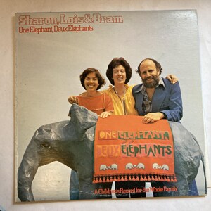 May include: Album cover for "One Elephant, Deux Éléphants" by Sharon, Lois & Bram. The image shows the musical trio with a paper mache elephant. The elephant has a red blanket with the album title. The text "A Children's Record for the Whole Family" is at the bottom.