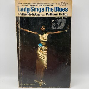 May include: Vintage paperback book titled "Lady Sings The Blues" by Billie Holiday with William Dufty. The cover features a photo of a woman in a gold dress with her arms outstretched. The book has a blue and white title and the text "WITH EXCLUSIVE PHOTOS".