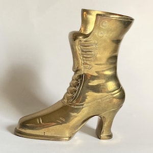 May include: A vintage brass boot sculpture with a high heel and intricate detailing. The boot features a lace-up design and a curved top, showcasing a classic, antique aesthetic. The golden color of the brass adds a touch of elegance.