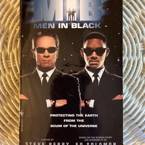 Peut inclure: Un livre de poche intitul&eacute; "Men in Black" avec deux hommes en costume noir et lunettes de soleil sur la couverture. Le titre du livre est au-dessus de l'image des deux hommes. Le texte dit "Prot&eacute;ger la Terre de la racaille de l'univers".