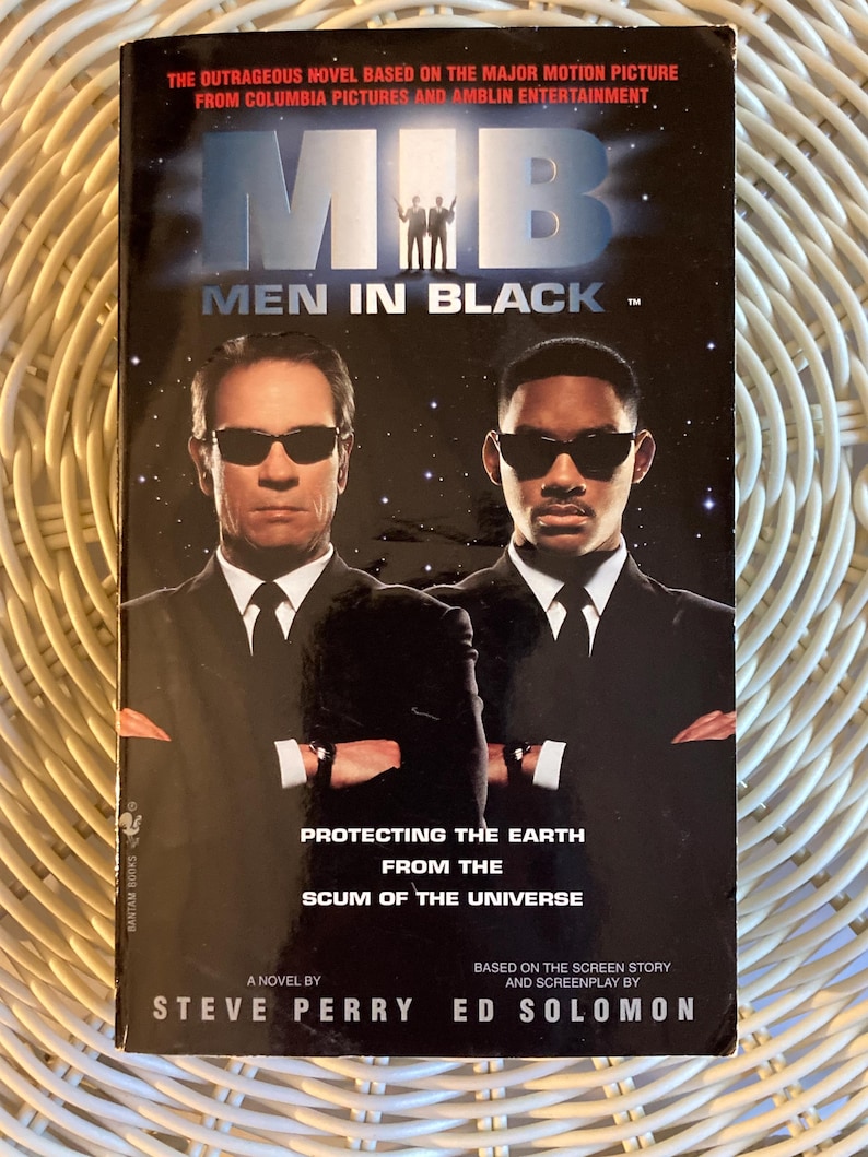 Peut inclure: Un livre de poche intitul&eacute; "Men in Black" avec deux hommes en costume noir et lunettes de soleil sur la couverture. Le titre du livre est au-dessus de l'image des deux hommes. Le texte dit "Prot&eacute;ger la Terre de la racaille de l'univers".