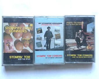 May include: Three vintage cassette tapes featuring album art for Stompin' Tom Connors. The tapes have different album titles, including "Bud the Spud" and "My Stompin' Grounds." The album covers feature images of the artist and other graphics.