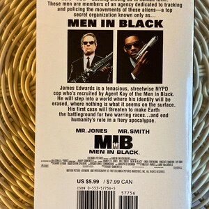Peut inclure: Une cassette VHS du film "Men in Black" avec deux hommes en costume noir tenant des armes. Le titre "Men in Black" est bien visible. Le dos de la cassette contient du texte d&eacute;crivant l'intrigue et les acteurs du film.