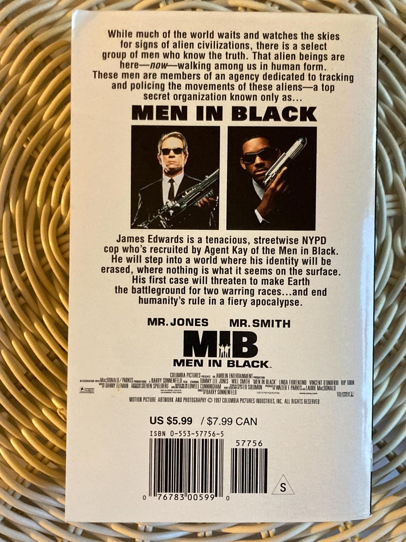 Peut inclure: Une cassette VHS du film "Men in Black" avec deux hommes en costume noir tenant des armes. Le titre "Men in Black" est bien visible. Le dos de la cassette contient du texte d&eacute;crivant l'intrigue et les acteurs du film.