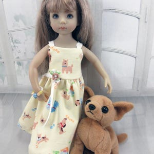 May include: A doll with long light brown hair wearing a pale yellow sleeveless dress featuring a repeating pattern of small dogs and the text "Ahooc". The doll has a small beaded bracelet and a flower accessory in its hair, standing next to a brown plush dog.